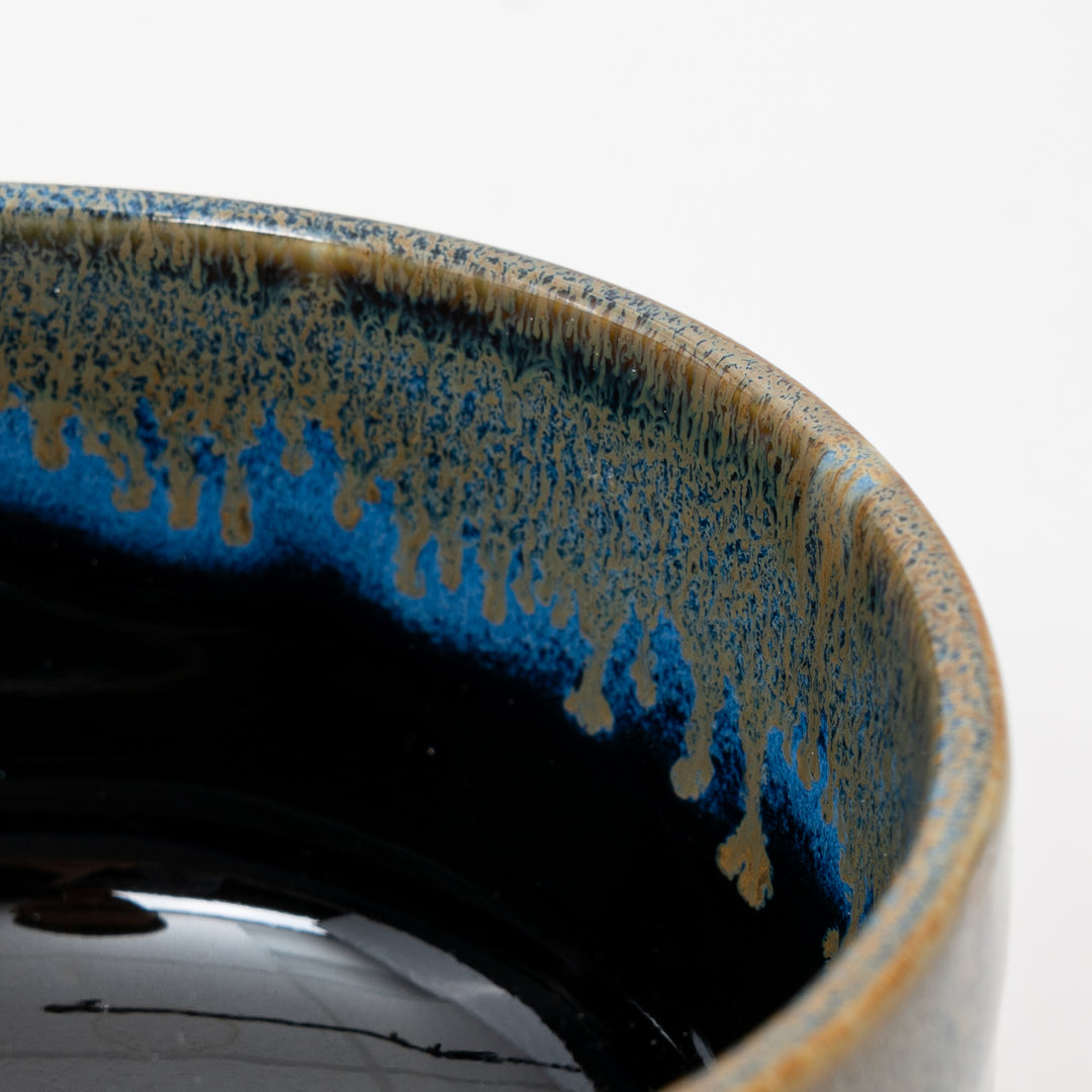 A close-up side view of a Japanese ceramic bowl, showing the beautiful contrast between the black body and the flowing blue drip glaze.