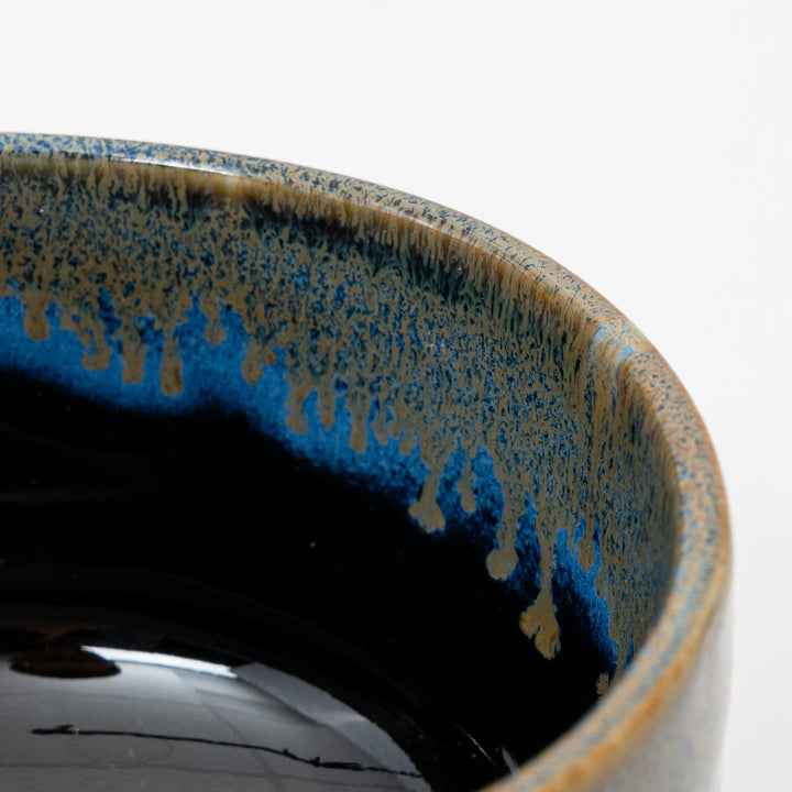 A close-up side view of a Japanese ceramic bowl, showing the beautiful contrast between the black body and the flowing blue drip glaze.