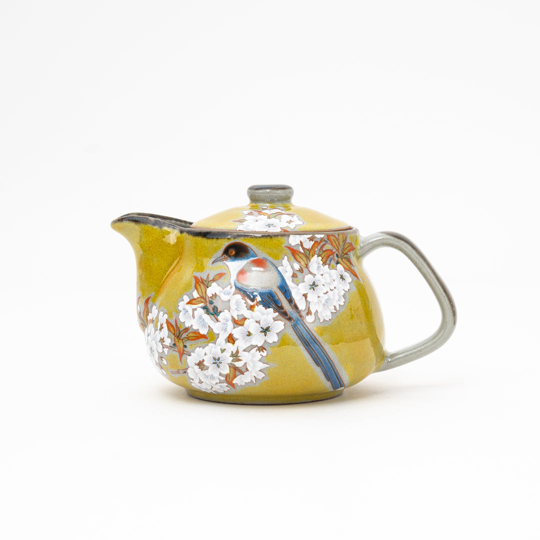 A yellow ceramic teapot with a floral and bird pattern, likely representing a cherry blossom design, on a plain background.