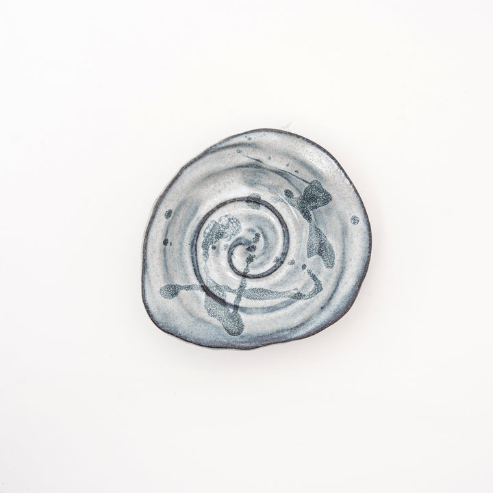 A top-down view of a small, irregularly shaped Japanese ceramic dish, showing a bold black spiral and splatter pattern.