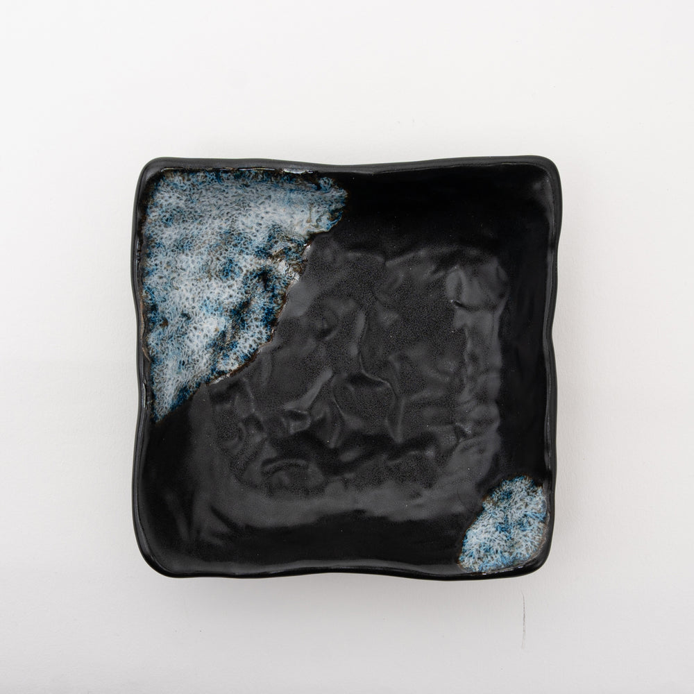 A close-up, angled view of the black ceramic bowl, showing the rich texture of the black surface and the glossy, speckled "Melted Snow" glaze.