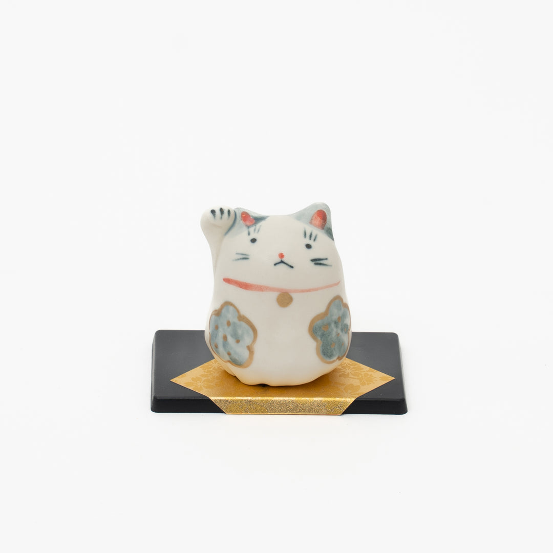  A white and grey Japanese Mino Yaki Yakushigama Maneki Neko lucky cat figurine with a red collar and bell, sitting on a black base with gold accents, against a white background.