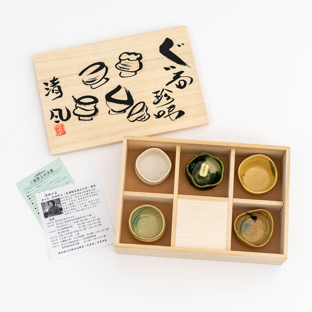 A set of handmade sake cups with different colors and textures, presented in a wooden box with Japanese characters printed on the lid.