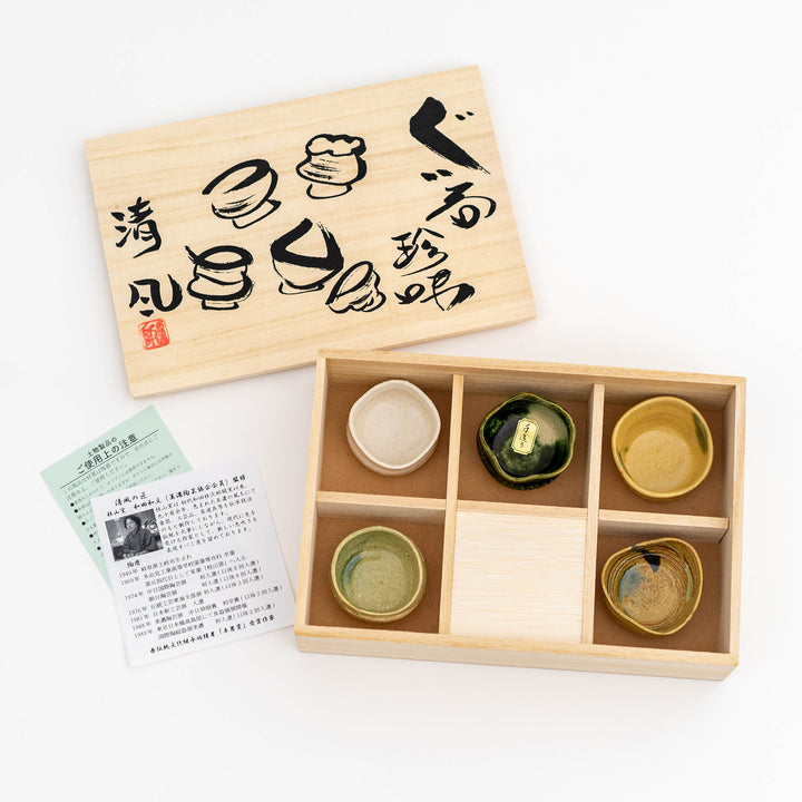 A set of handmade sake cups with different colors and textures, presented in a wooden box with Japanese characters printed on the lid.